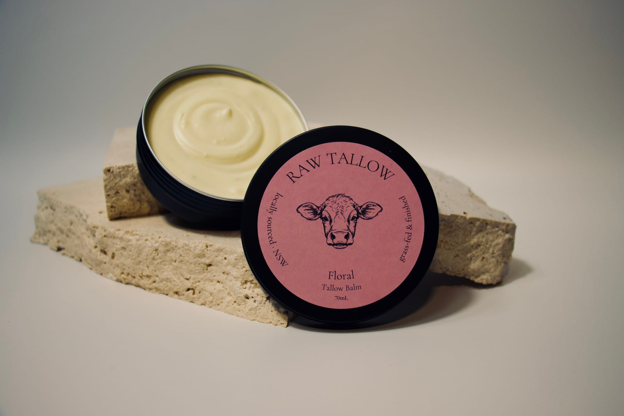 Black amber jar of Raw Tallow Floral Tallow Balm with soft pink circular label on travertine and dried florals